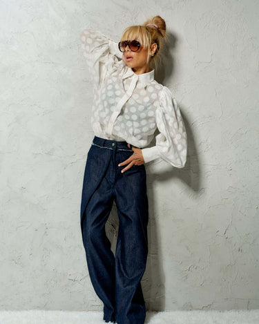 Model in a white sheer polka dot puff sleeve shirt and dark denim pants, showcasing a stylish, sophisticated look.