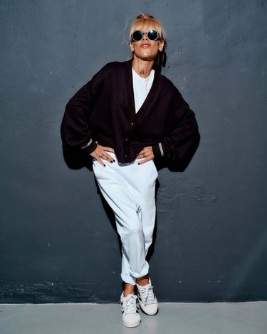 Woman posing in a black snap-button jacket over a white outfit, styled with sunglasses and sneakers.