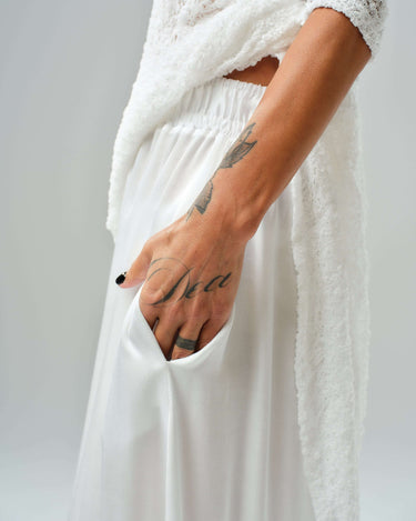 Close-up of a model's hand in a white maxi skirt pocket, showcasing a tattoo and layered tunic.