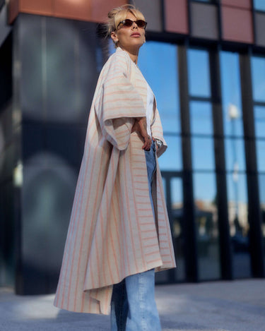 Model showcasing a striped linen-cotton kimono, styled with wide-leg jeans and sunglasses, against a modern urban backdrop.