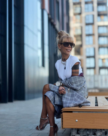 Woman in oversized checked blazer and sunglasses, sitting on a bench outdoors with a relaxed smile.