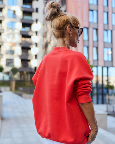 Woman in bold red oversized sweatshirt, showcasing urban streetwear style against a modern city backdrop.