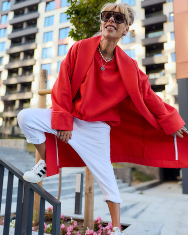 Model wearing a red oversized coat with large pockets, styled casually with white pants and sunglasses, showcasing a relaxed silhouette.