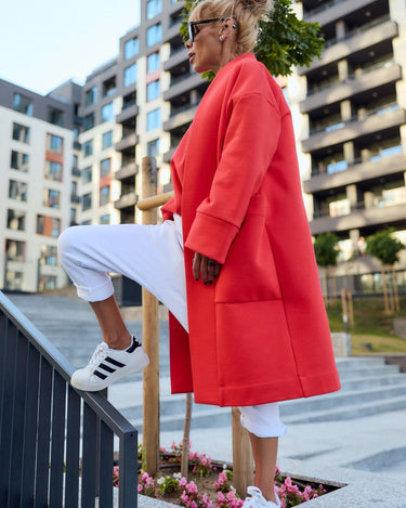 Stylish woman in a red oversized coat, white pants, and sneakers, posing outdoors for a trendy urban look.