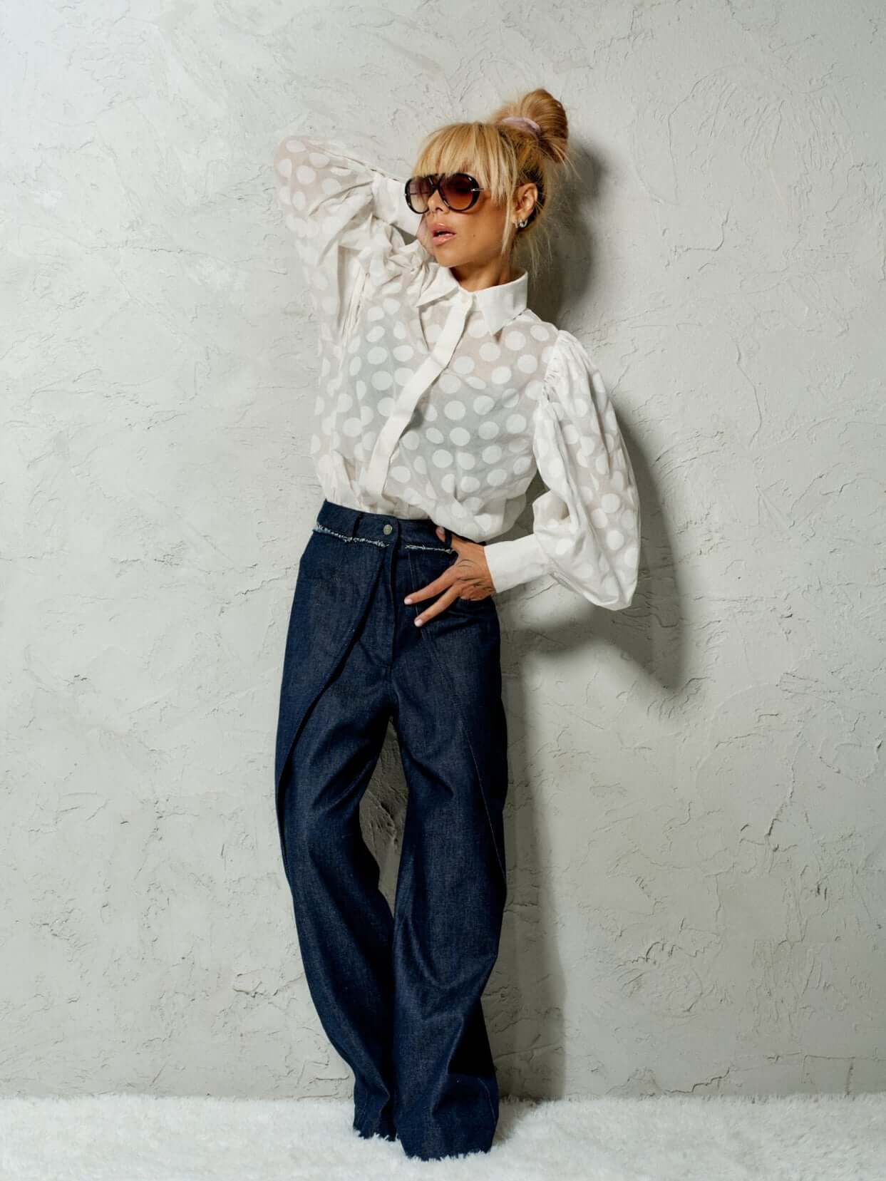 Model in a white sheer polka dot puff sleeve shirt and dark denim pants, showcasing a stylish, sophisticated look.