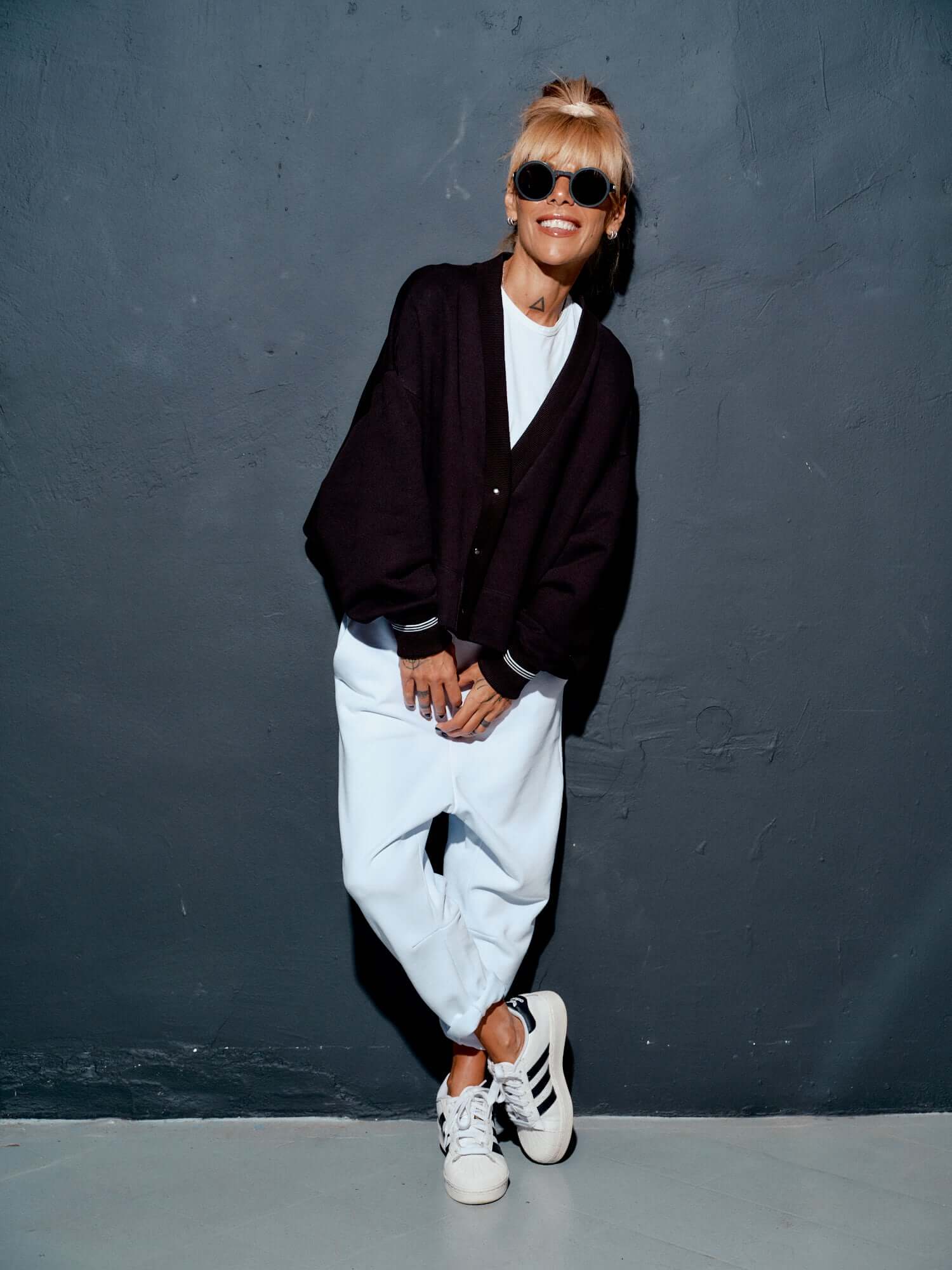 Woman wearing a black snap-button jacket over a white shirt and light trousers, posing against a gray wall.