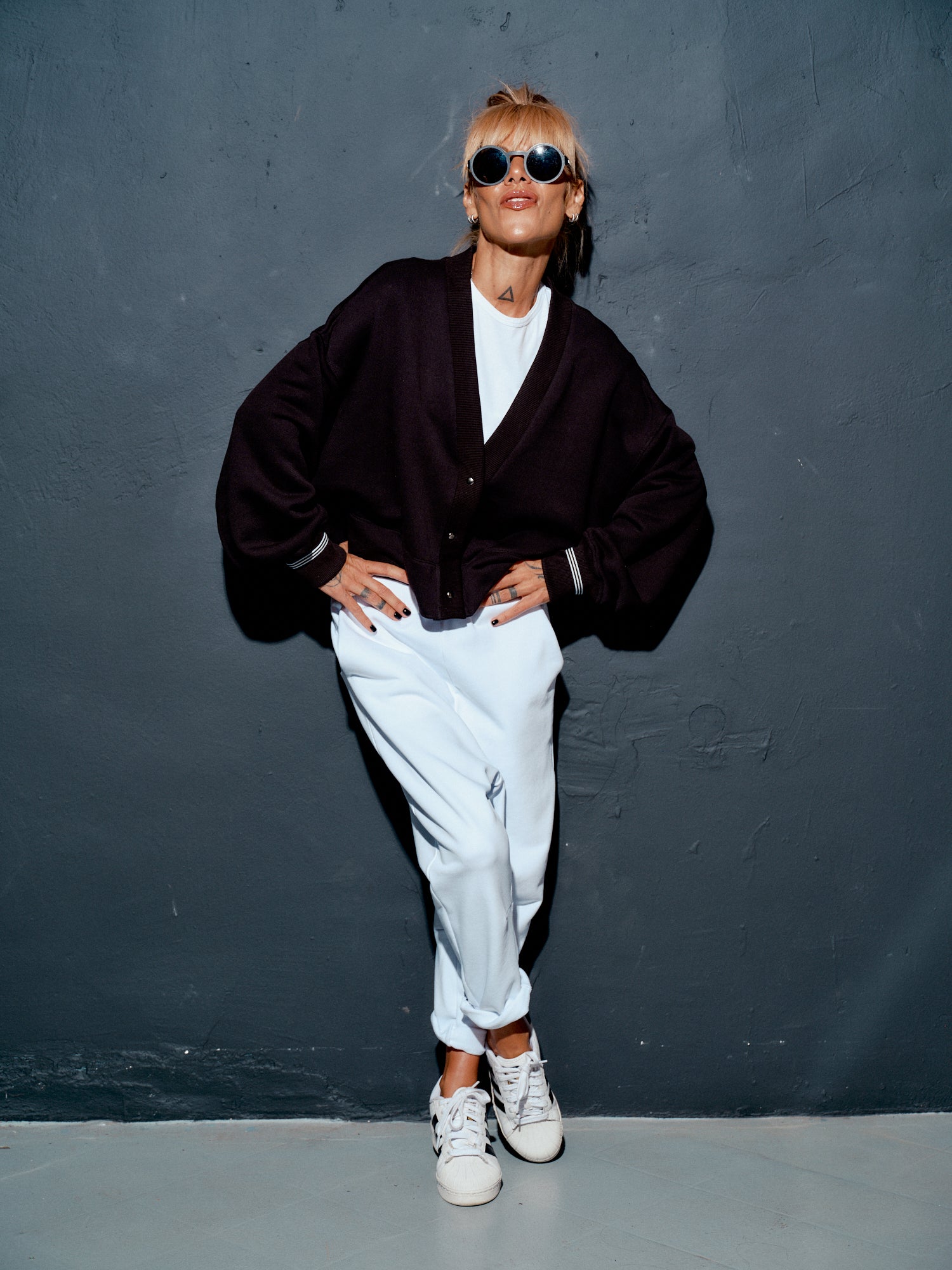 Woman posing in a black snap-button jacket over a white outfit, styled with sunglasses and sneakers.
