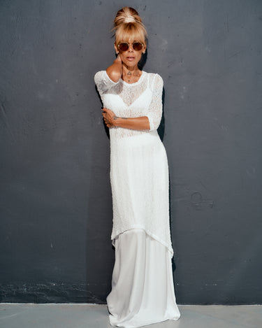 Model wearing a white maxi co-ord set with a sheer tunic and flowing elastic waist skirt against a gray wall.