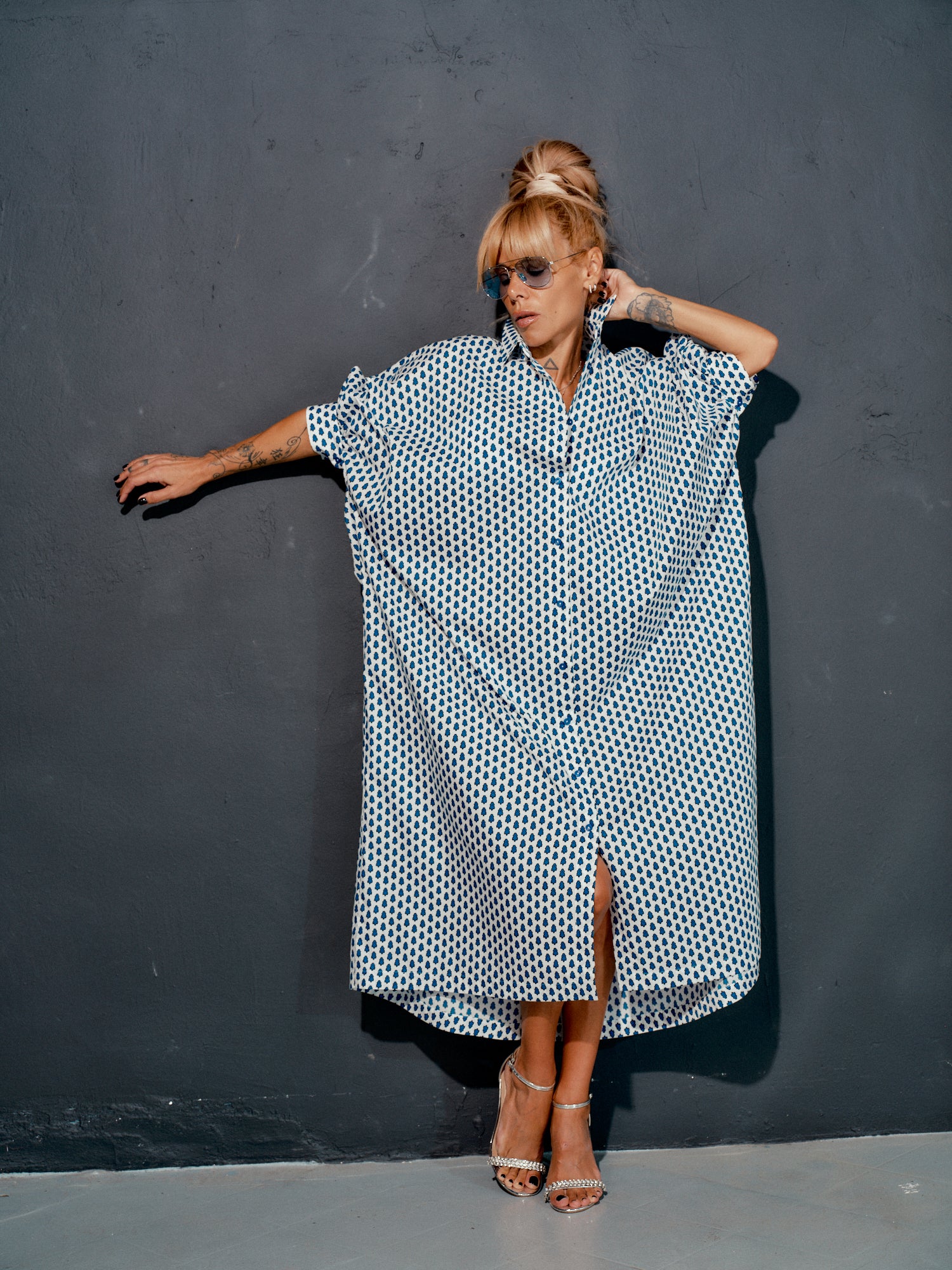 Woman wearing an oversized shirt dress with a blue micro floral print, styled with sunglasses against a dark wall.