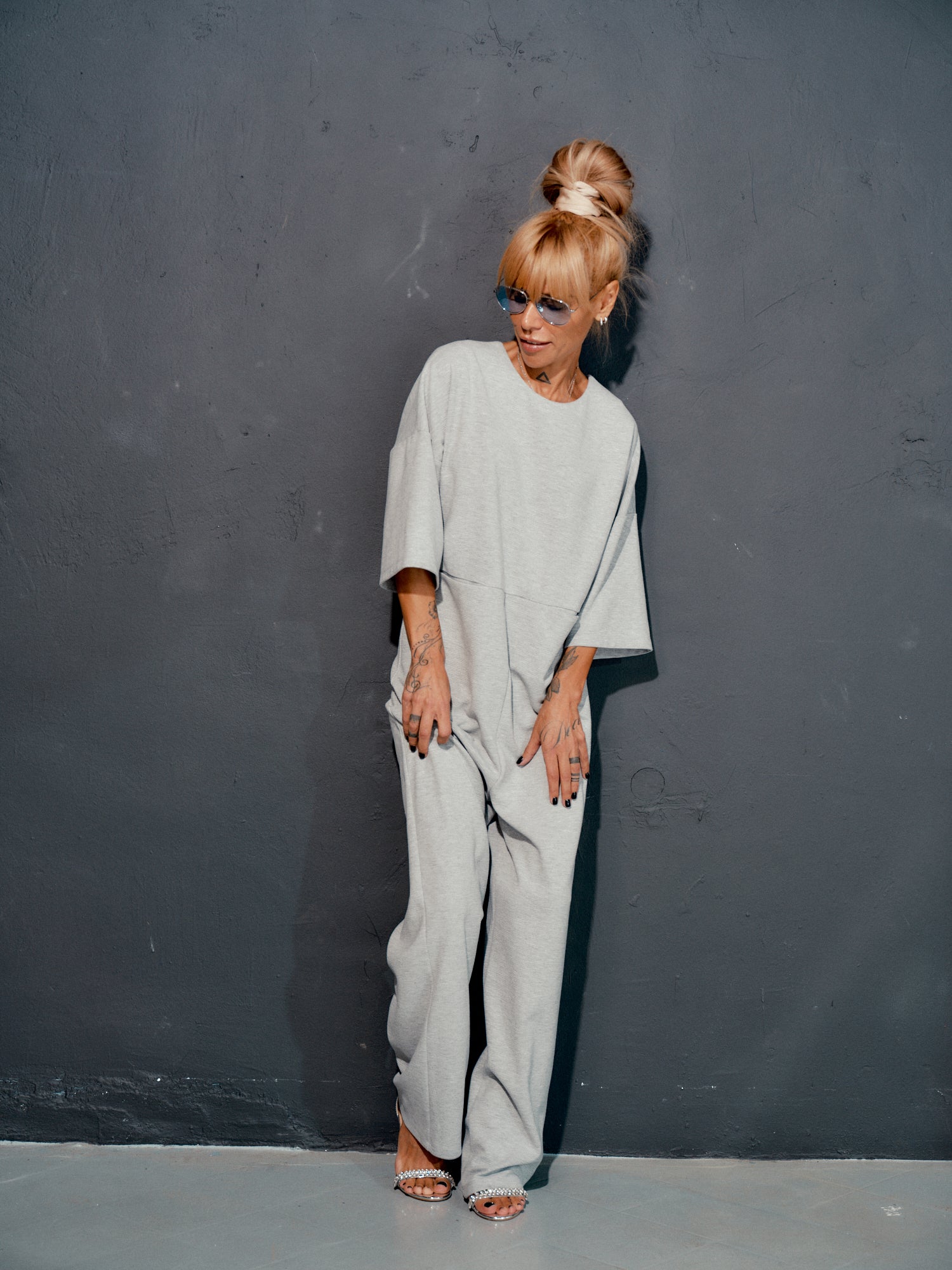 Grey wide-leg jumpsuit with open back, featuring cropped sleeves and side pockets, modeled against a dark wall.