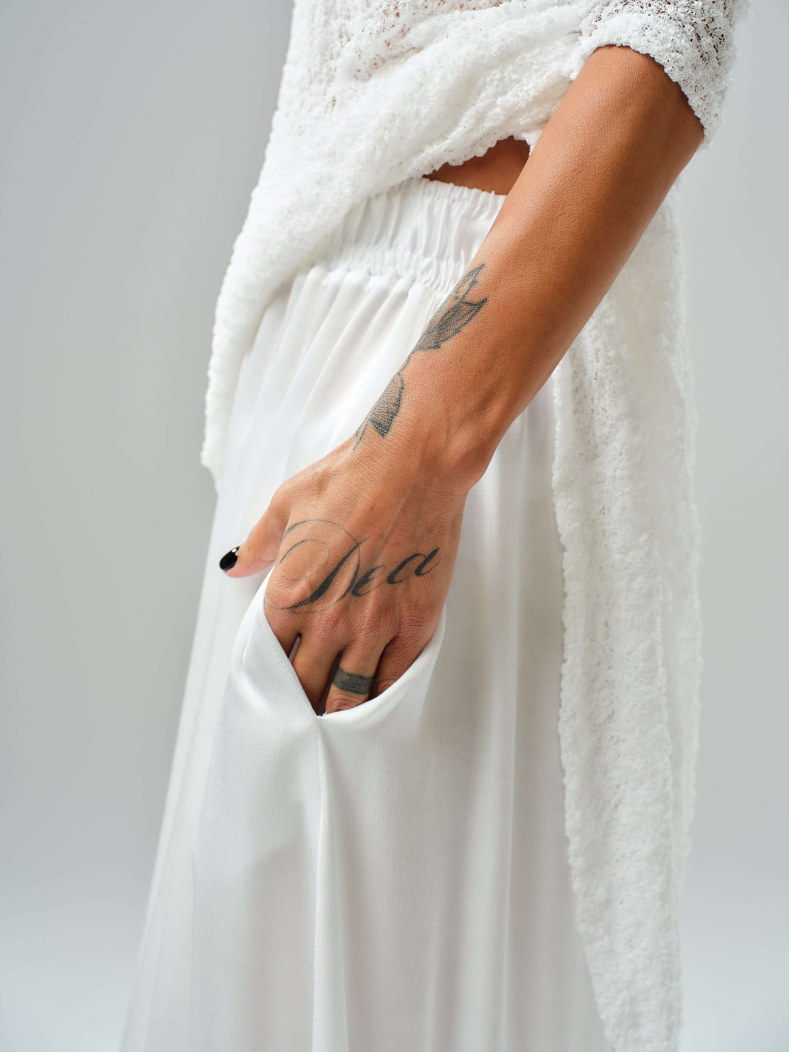 Close-up of a model's hand in a white maxi skirt pocket, showcasing a tattoo and layered tunic.