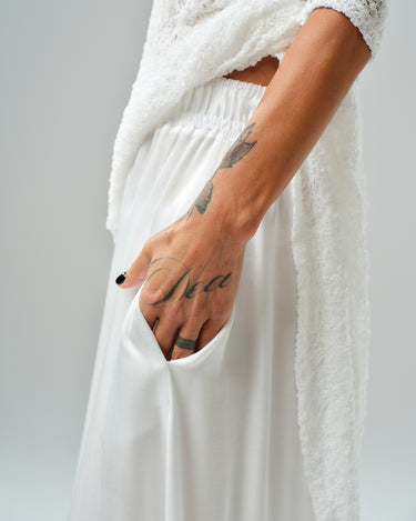 Close-up of a model's hand in a white maxi skirt pocket, showcasing a tattoo and layered tunic.