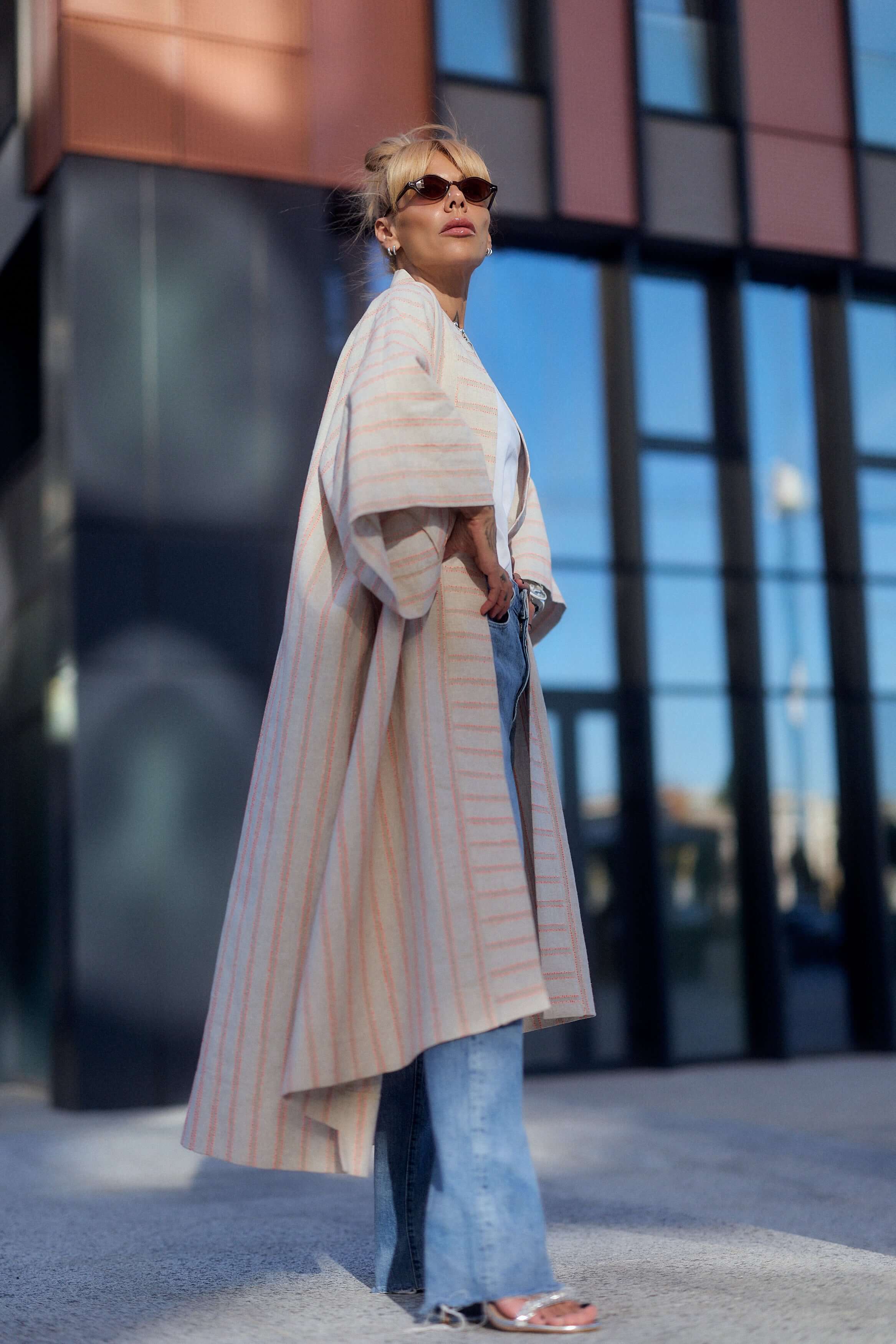 Model showcasing a striped linen-cotton kimono, styled with wide-leg jeans and sunglasses, against a modern urban backdrop.