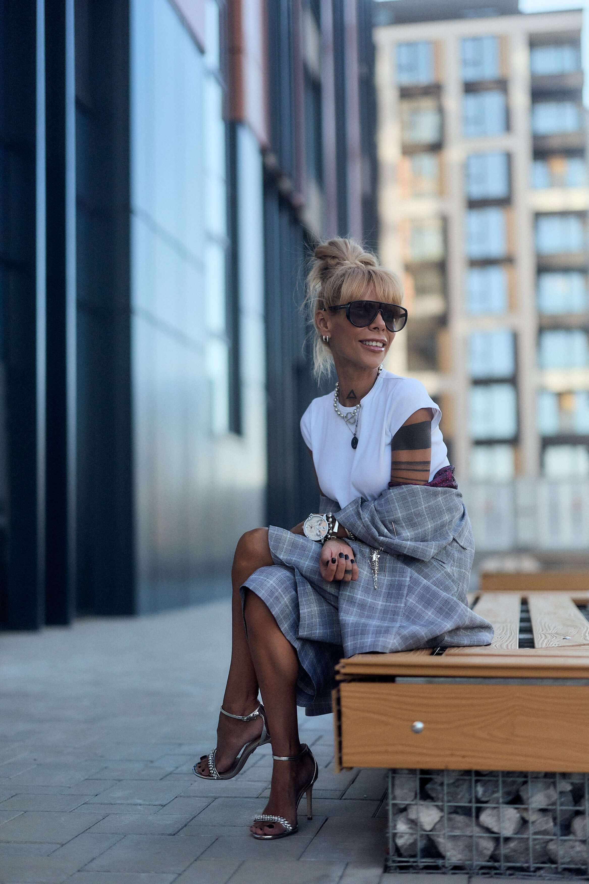 Woman in oversized checked blazer and sunglasses, sitting on a bench outdoors with a relaxed smile.