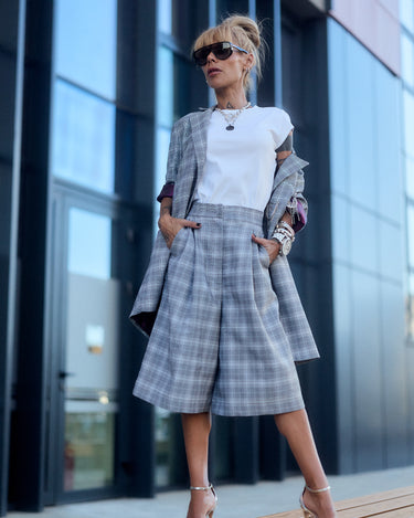 Woman in a light grey checked blazer and culotte set, posing confidently outdoors with stylish accessories.