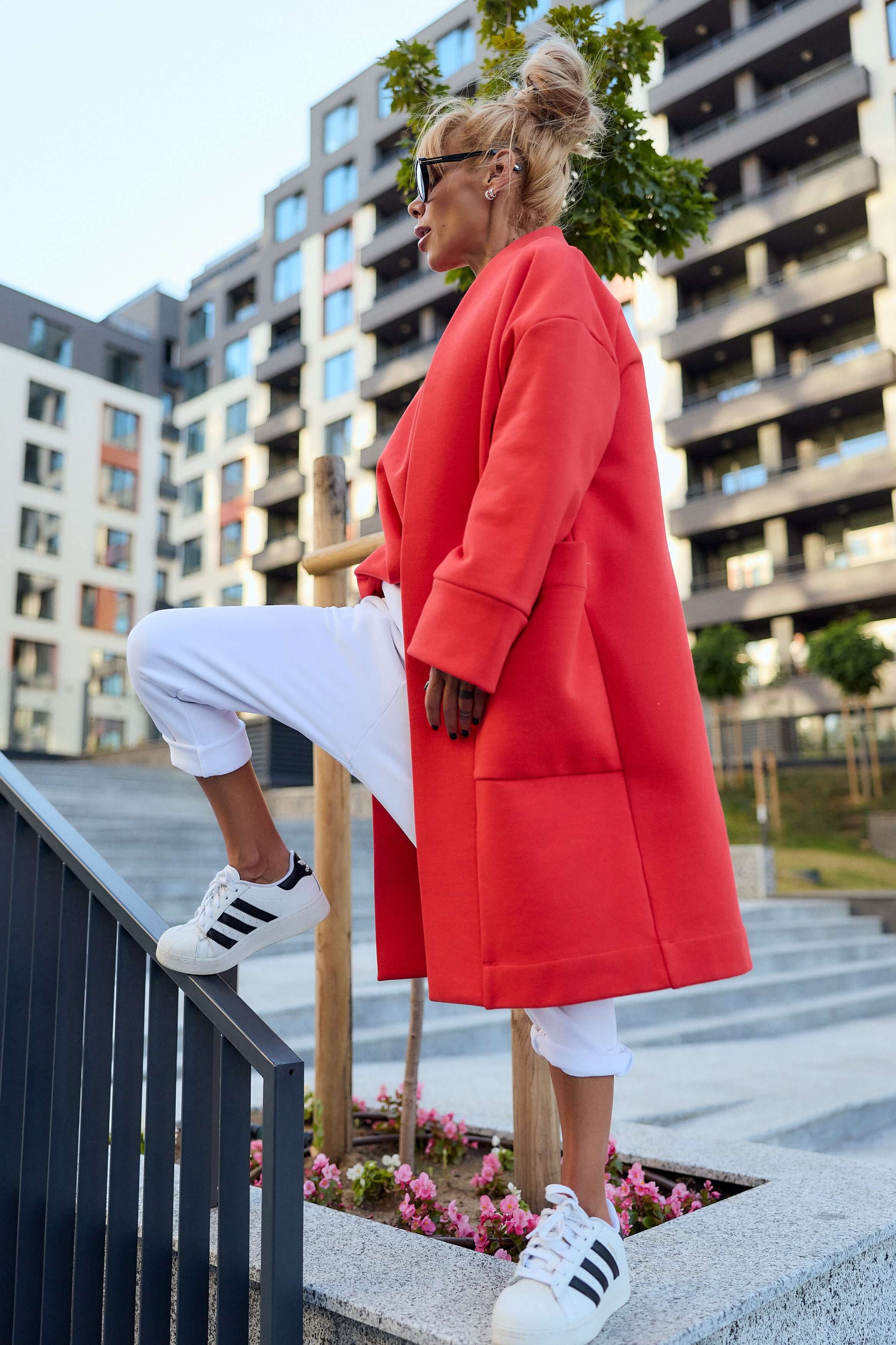 Stylish woman in a red oversized coat, white pants, and sneakers, posing outdoors for a trendy urban look.