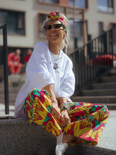 Person sitting on steps wearing a white hoodie and colorful pants with sunglasses and a headband.