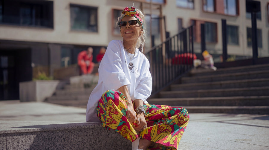 Person wearing a white jacket and colorful pants sitting on a ledge with a blurred urban background