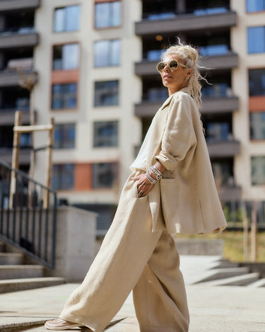 Person wearing a beige outfit walking on a city street with buildings in the background