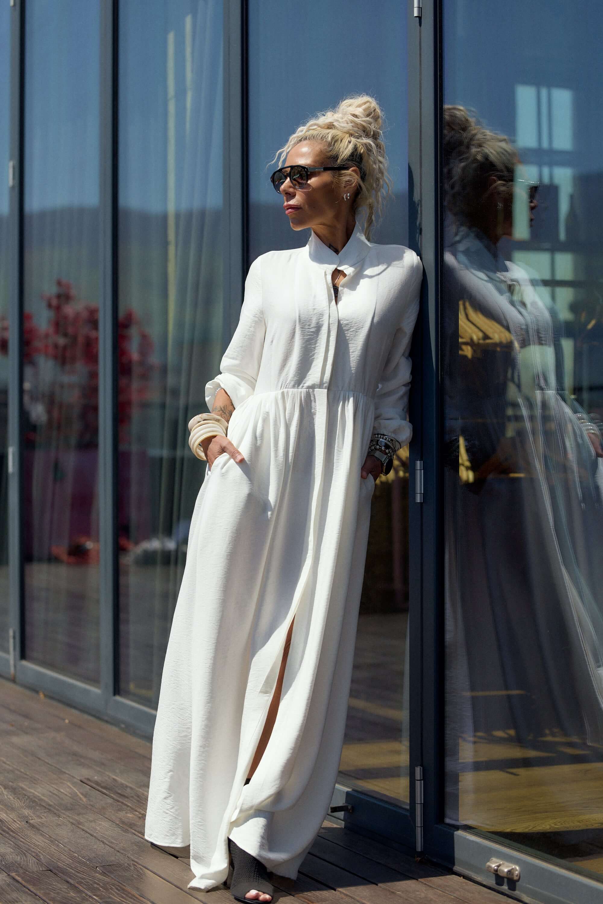 Flowing White Maxi Dress worn by a woman leaning against glass doors, showcasing its elegant style and flowing silhouette.
