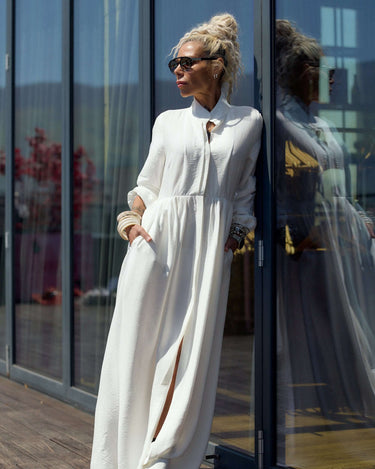 Flowing White Maxi Dress worn by a woman leaning against glass doors, showcasing its elegant style and flowing silhouette.