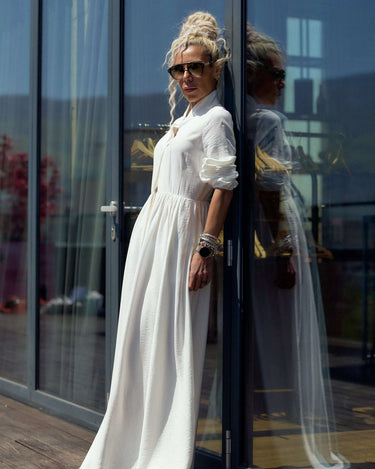 Model wearing a Flowing White Maxi Dress standing by a glass door, showcasing its elegant design and flowing fabric.