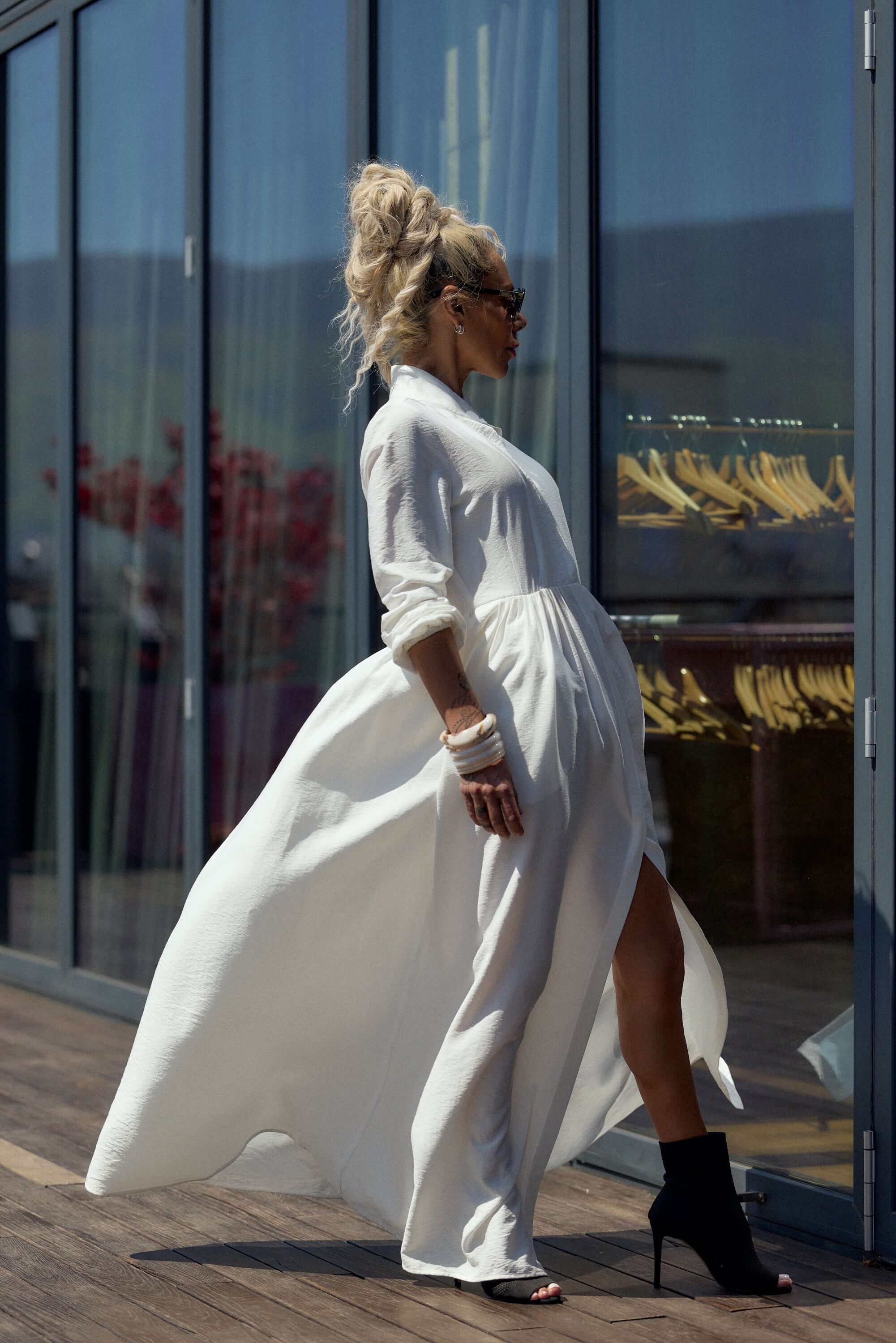 Woman in Flowing White Maxi Dress walking confidently near glass windows.