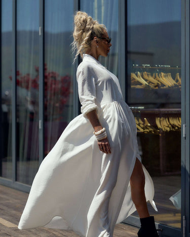 Woman in Flowing White Maxi Dress walking confidently near glass windows.