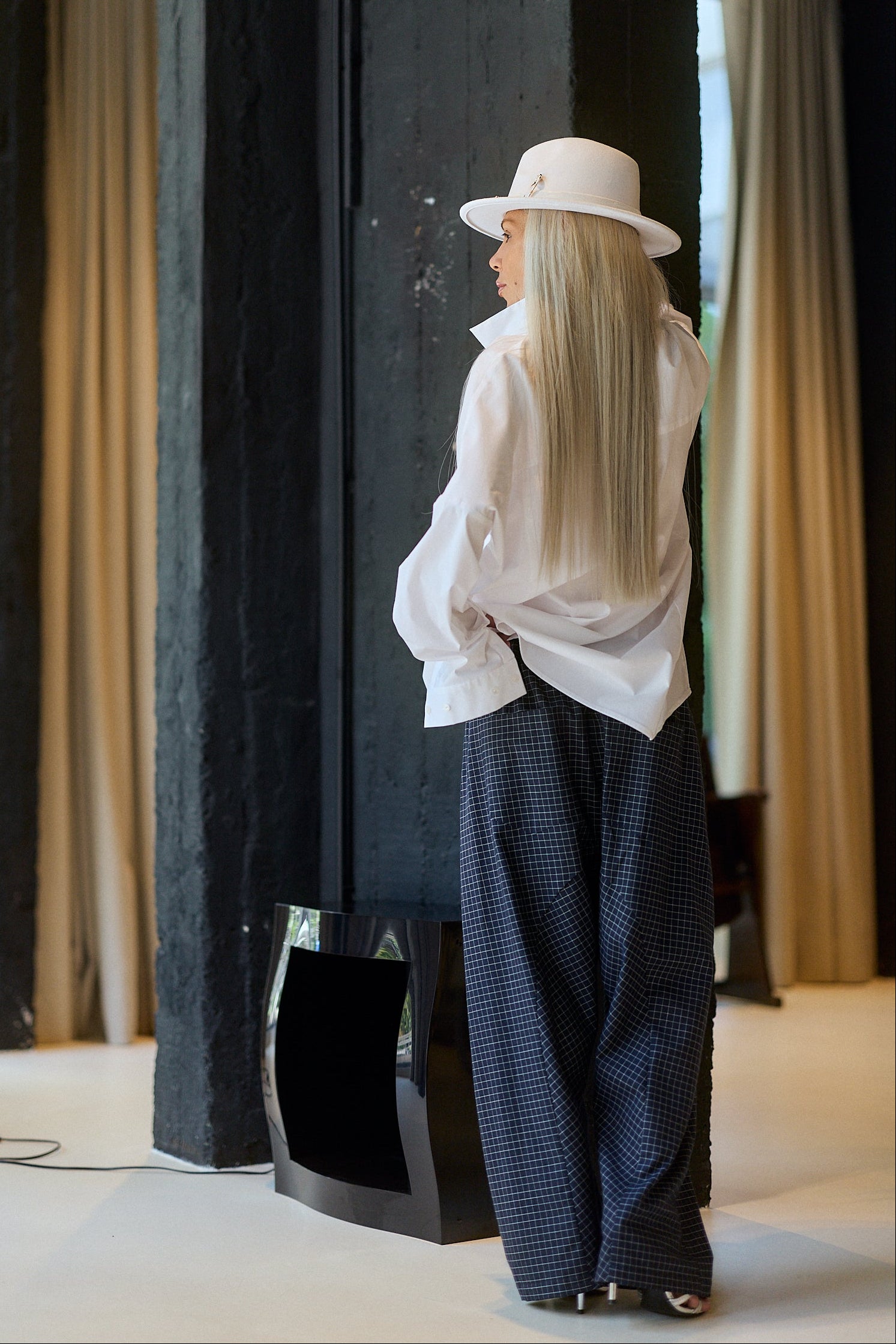 A woman with long blonde hair stands in profile wearing a white wide-brim hat, a loose white blouse, and dark blue gingham wide-leg trousers in a modern interior with dark columns and beige curtains.