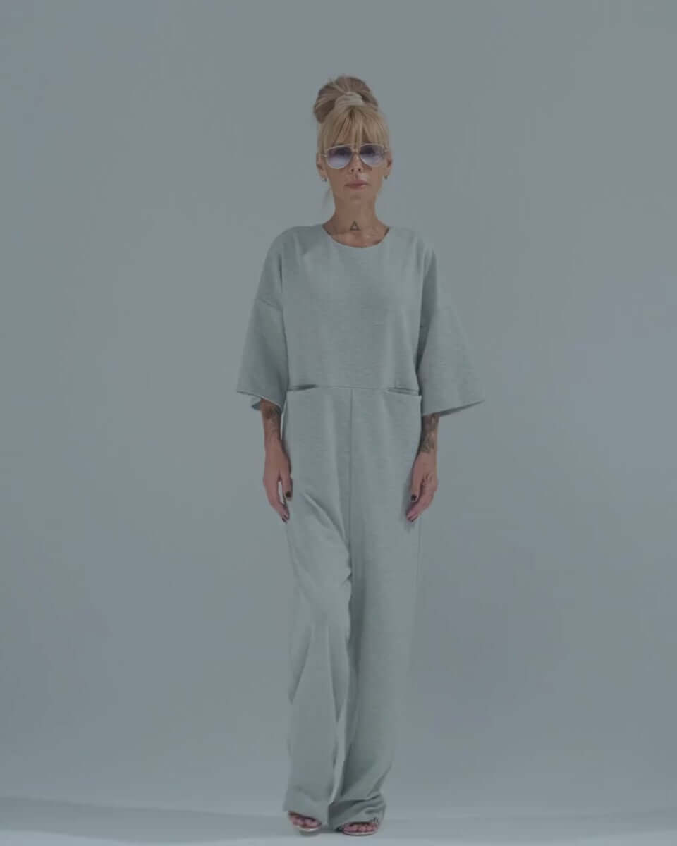 Stylish woman modeling a grey wide-leg jumpsuit with cropped sleeves and side pockets on a minimalist background.